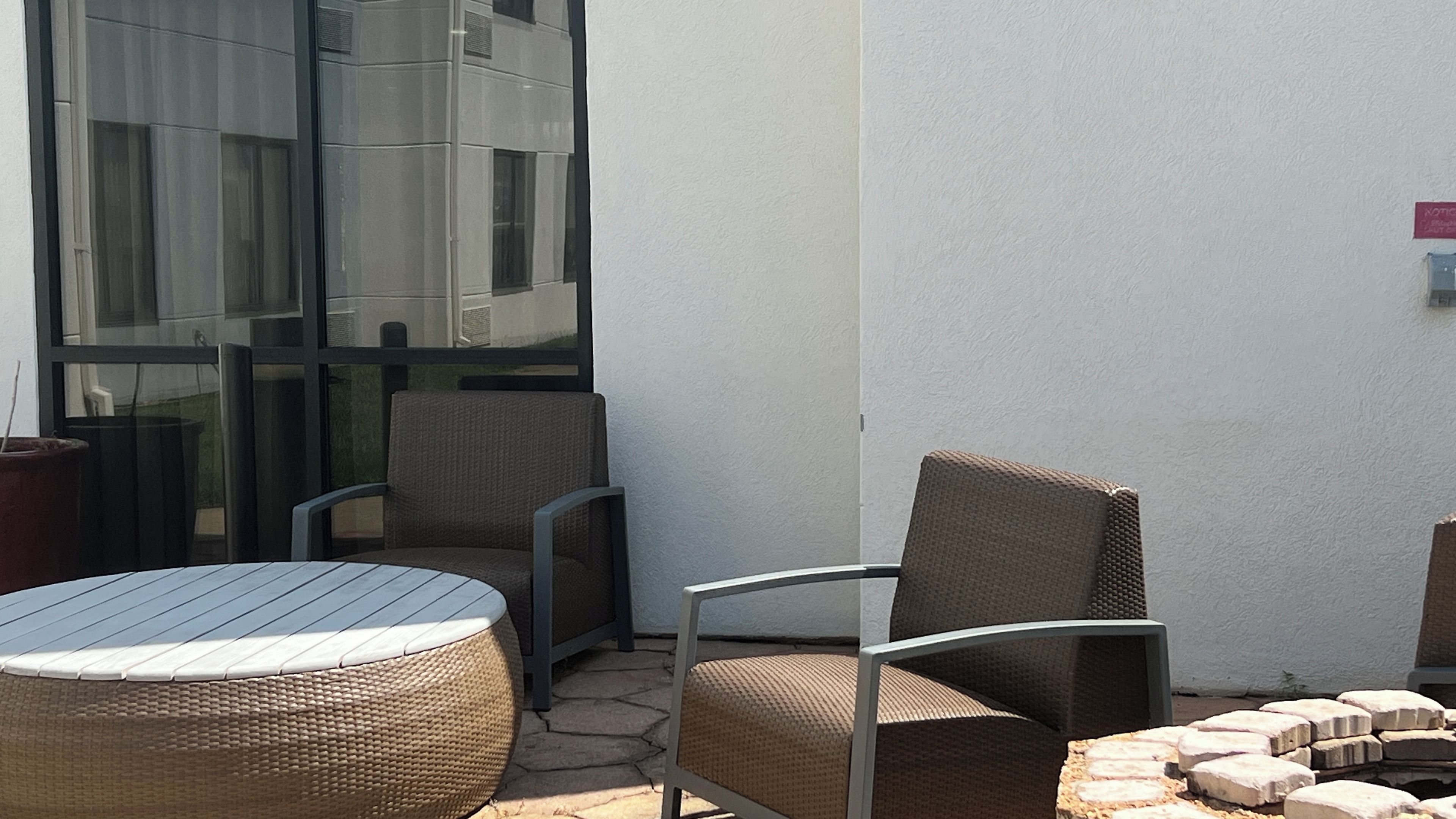 Uma área externa com mobiliário de vime e uma mesa de vidro, localizada ao lado de um edifício com grandes janelas.