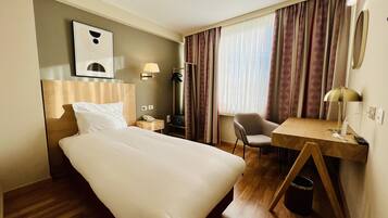 A hotel room with a bed, a desk, a chair, a lamp, a phone, and a window with curtains.