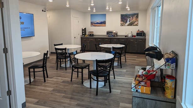 A modern kitchen with wooden flooring, white cabinets, and a dining area with round tables and chairs. - Henry's Fork Inn (St. Anthony)