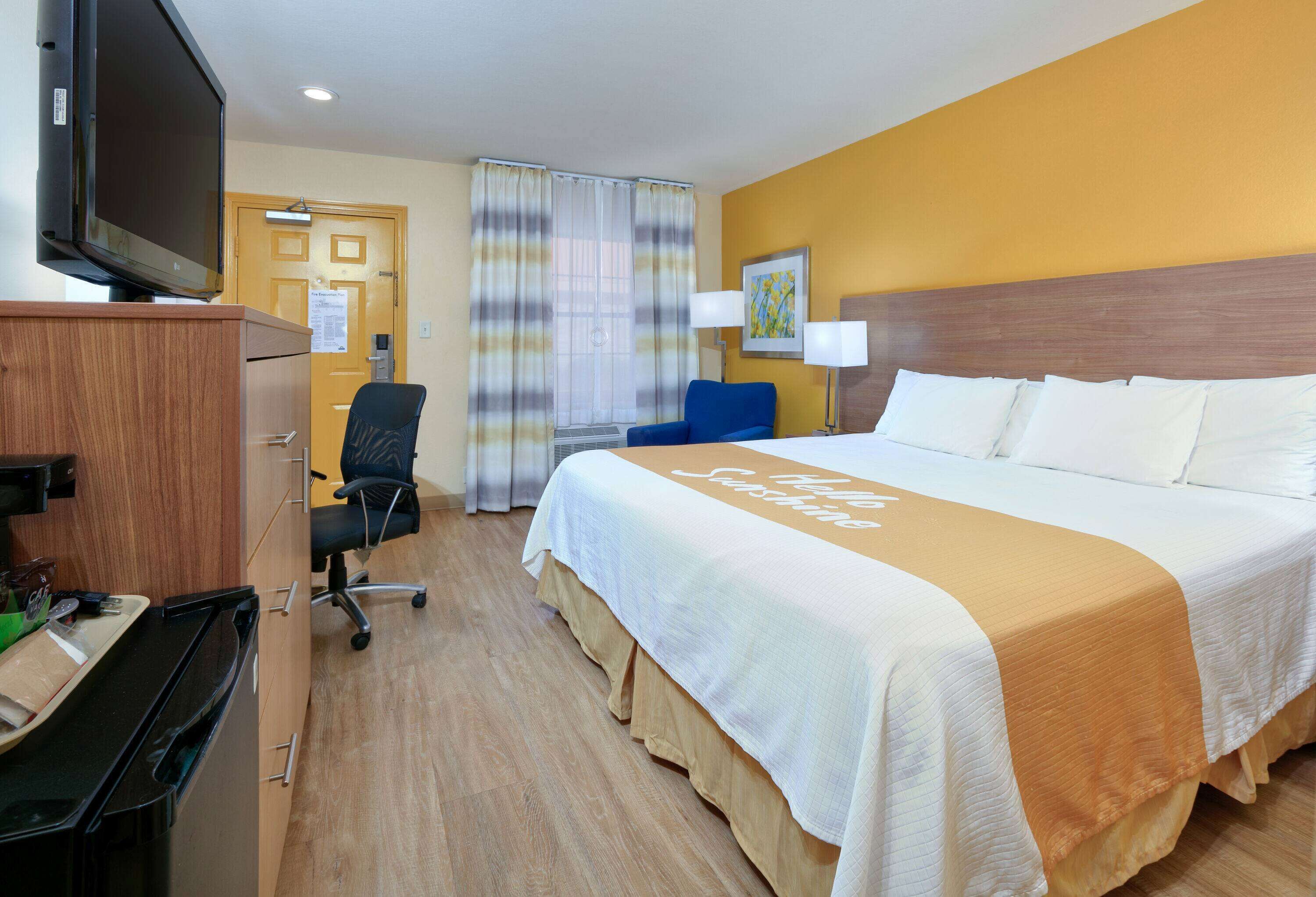 A hotel room with a bed, a desk with a television, a chair, and a desk lamp.