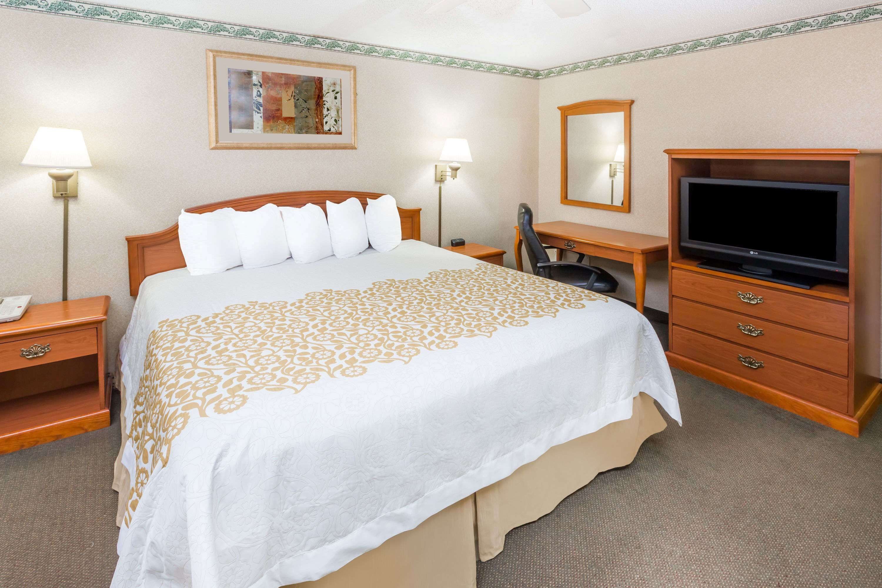 A hotel room with a bed, a desk with a TV, a chair, and a nightstand with a lamp.