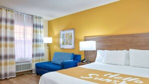 Ein Hotelzimmer mit einem Bett, einem blauen Sessel, zwei Nachttischlampen, einem gerahmten Bild an der Wand und einem Fenster mit Vorhängen.