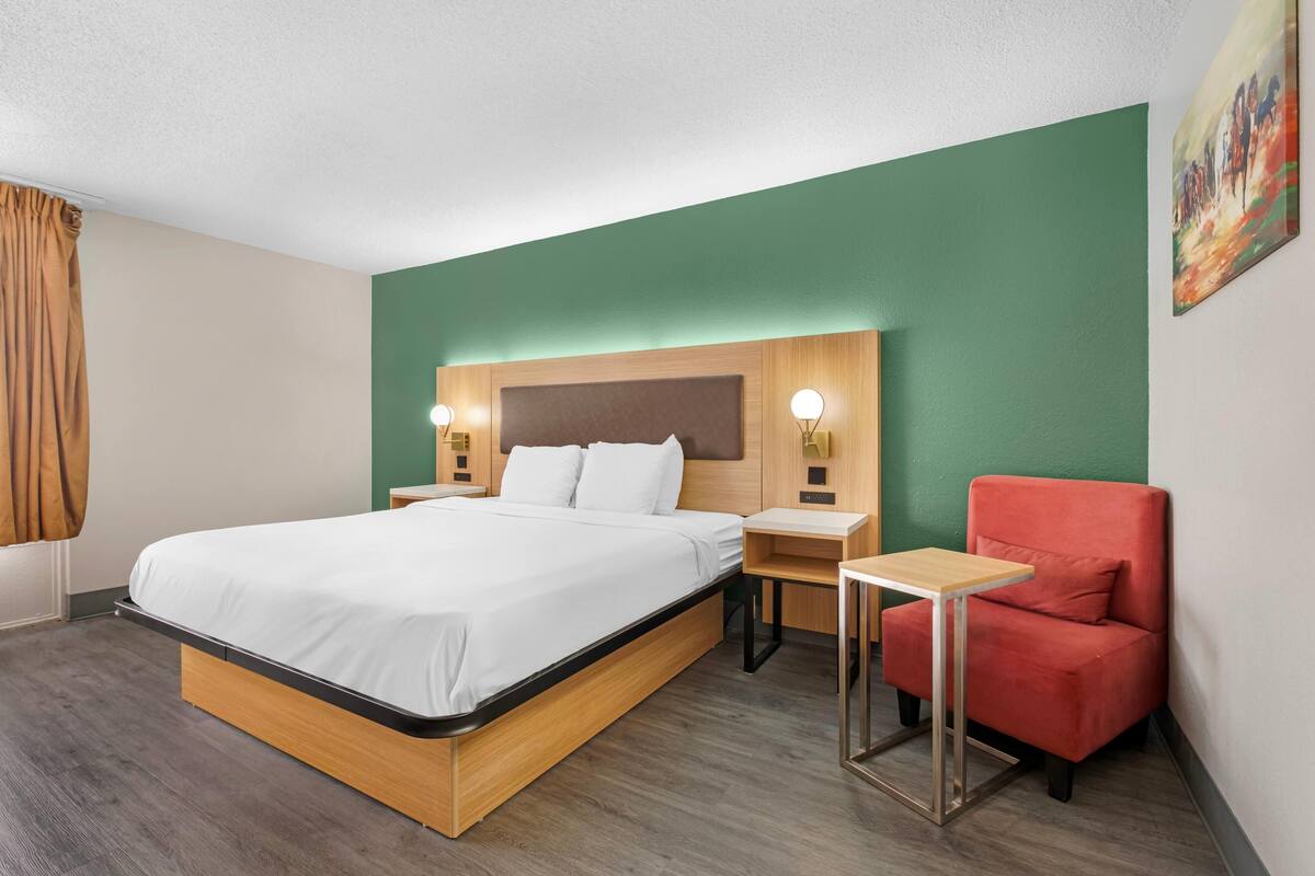 Une chambre d’hôtel avec un lit, une chaise rouge, une petite table et deux lampes de chevet.