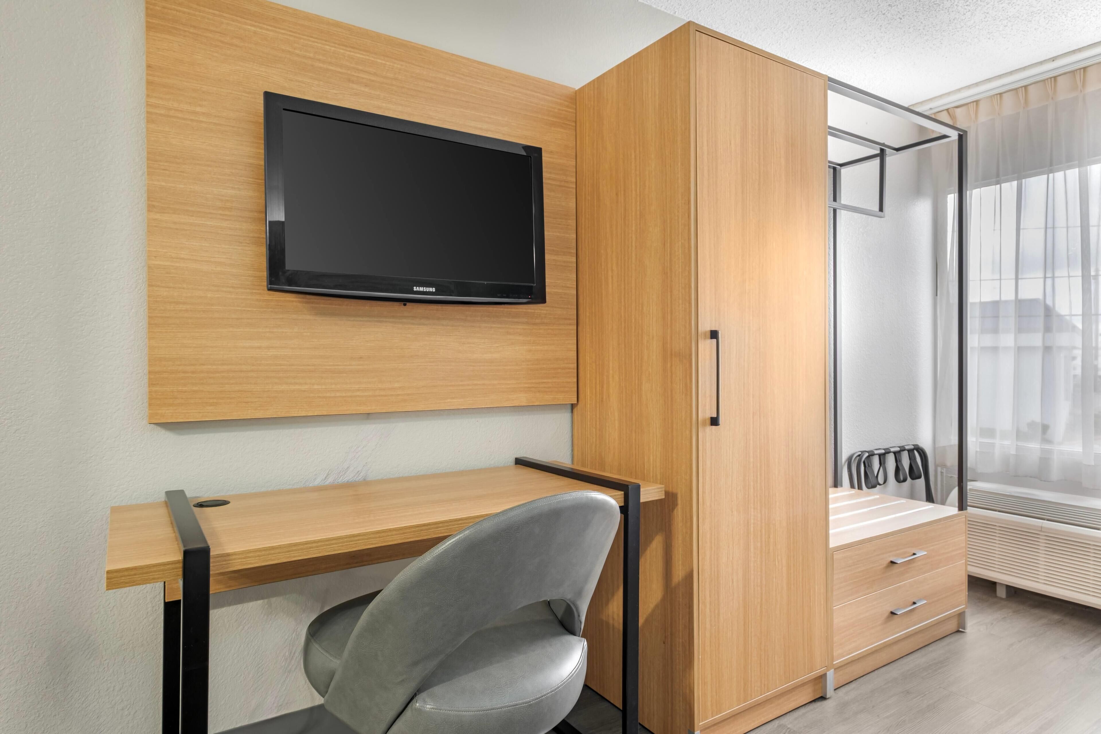 A modern room with a wooden desk, a grey chair, a TV mounted on the wall, and a wardrobe with a mirror.