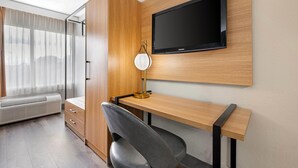 Habitación de hotel moderna con un escritorio de madera, un televisor de pantalla plana, una lámpara de escritorio, una silla, un armario y una ventana con cortinas traslúcidas.