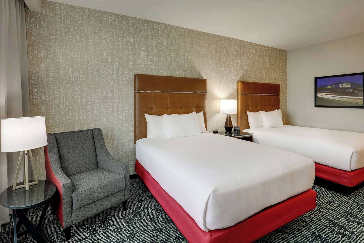 Ein Hotelzimmer mit zwei Betten, einem grauen Sessel, einem kleinen Tisch mit Lampe und einer Wand mit Muster.