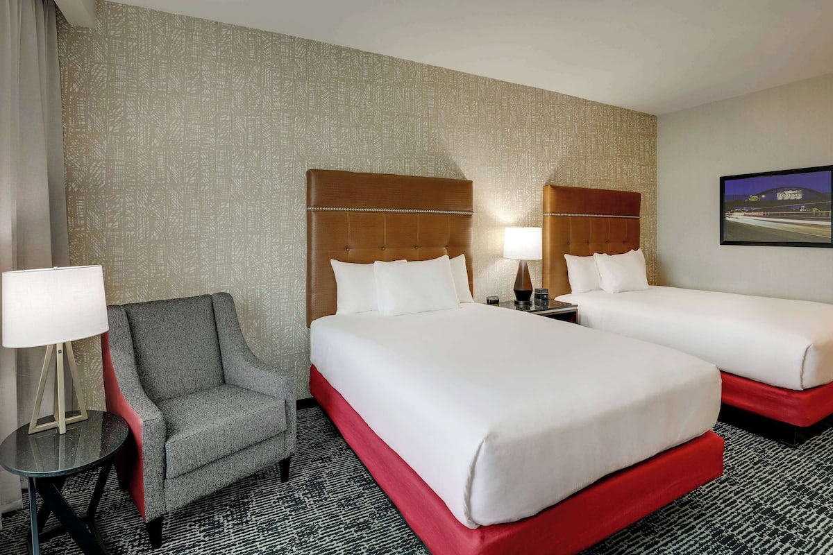 A hotel room with two beds, a grey armchair, a small table with a lamp, and a patterned wall.