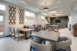 A modern dining area with multiple tables and chairs, a kitchen area with a flat-screen TV, and decorative wall panels.