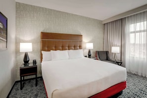 A hotel room with a large bed, two bedside tables with lamps, a chair, and a view of the city through the window.