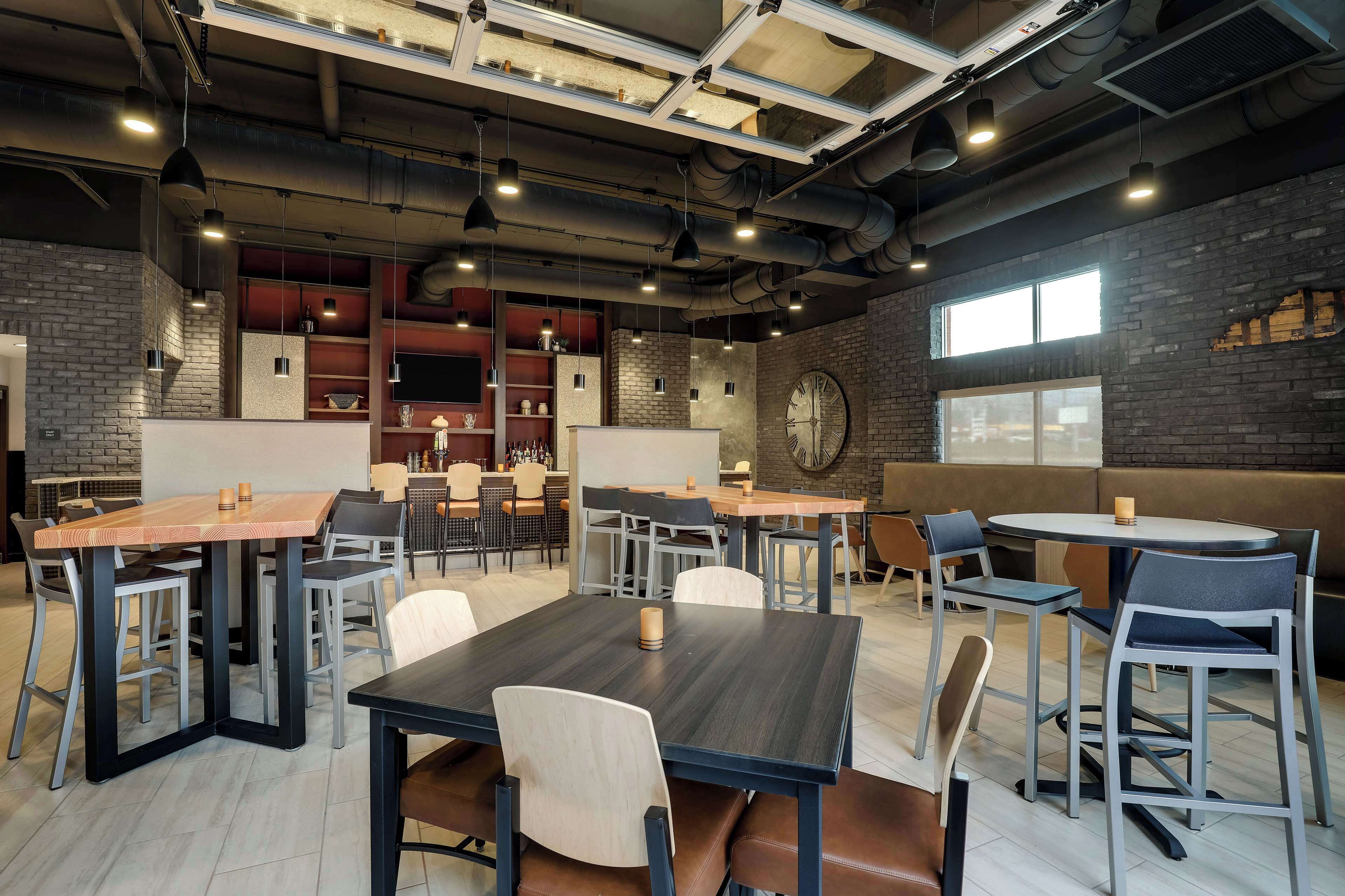 Uma sala de jantar moderna com várias mesas e cadeiras, dutos aparentes e paredes de tijolo.