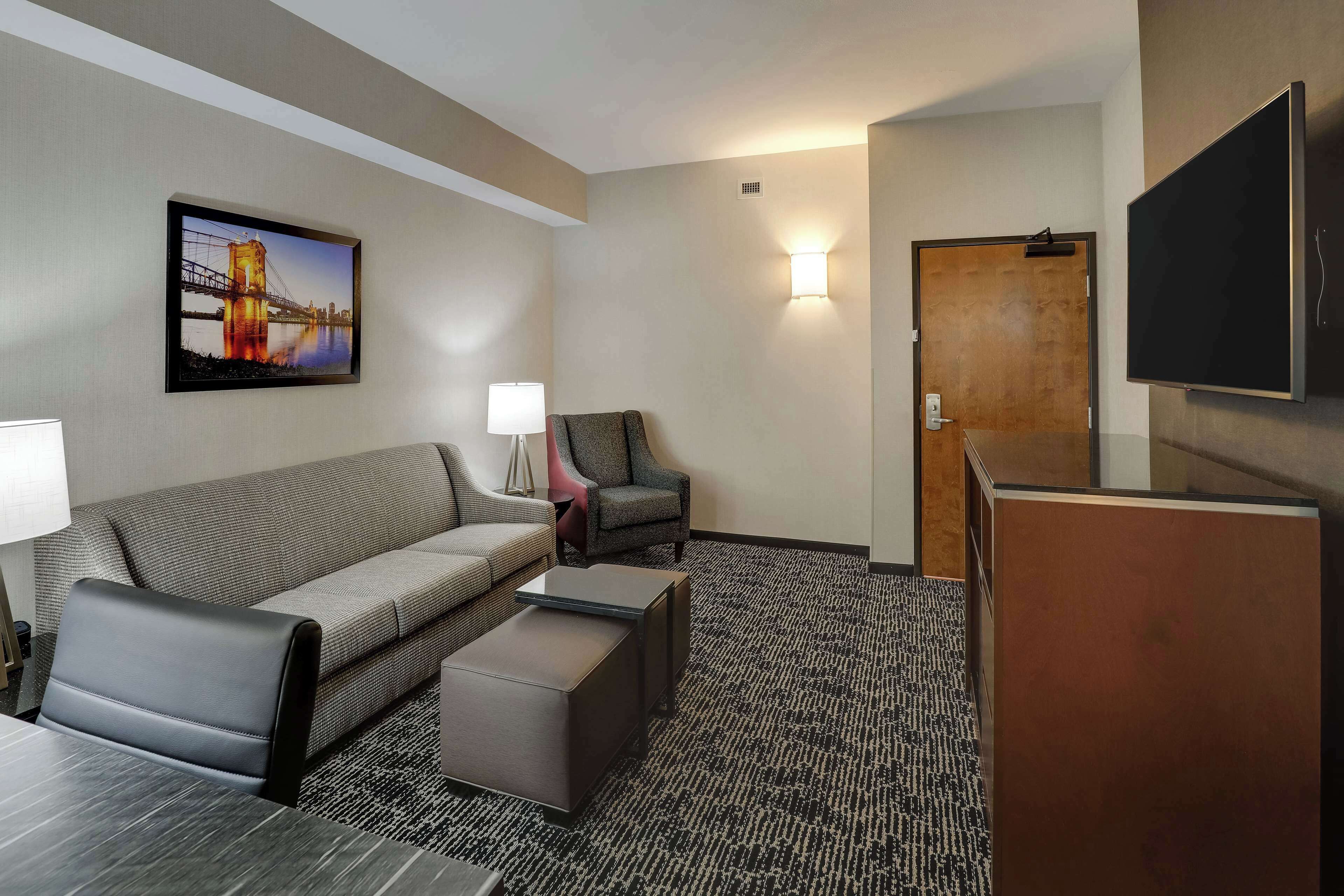 Une chambre d’hôtel équipée d’un canapé, d’un fauteuil, d’un pouf, d’un téléviseur à écran plat et d’un tableau représentant un pont.