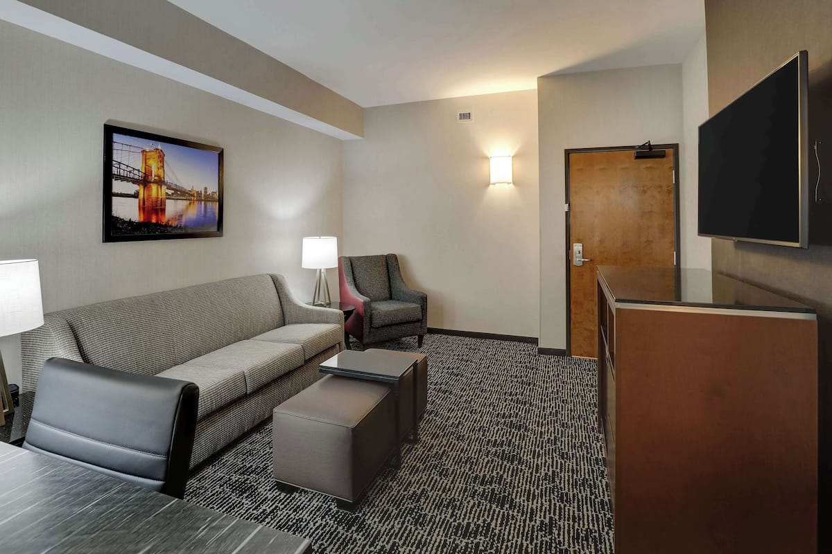 Ein Hotelzimmer mit Sofa, Sessel, Hocker, Flachbildfernseher und einem Gemälde einer Brücke.