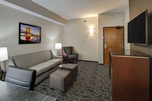A hotel room with a sofa, armchair, ottoman, flat-screen TV, and a painting of a bridge.