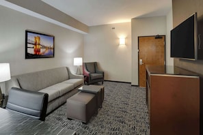 Habitación de hotel con sofá, sillón, otoman, televisor de pantalla plana y un cuadro de un puente.