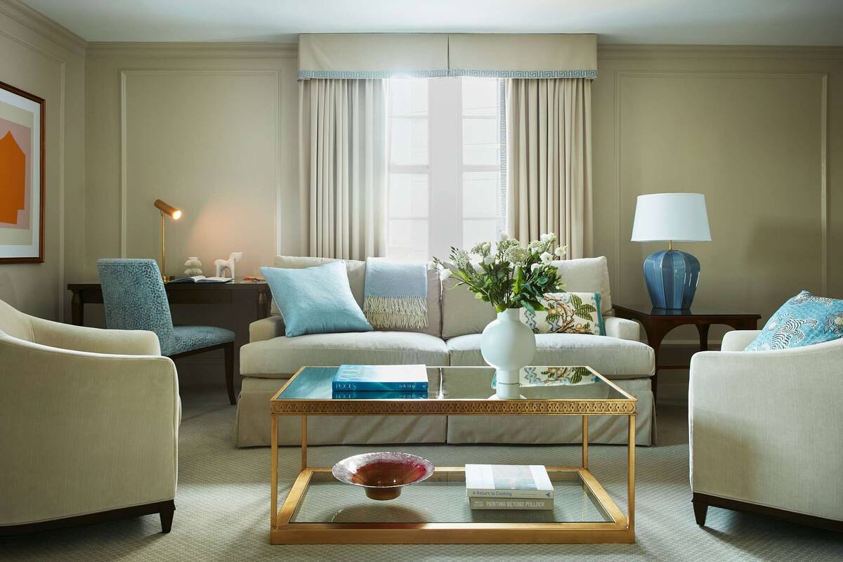 A well-lit living room with a sofa set, a coffee table, and two armchairs. There is a lamp on a side table, a vase with flowers on the coffee table, and a framed picture on the wall.