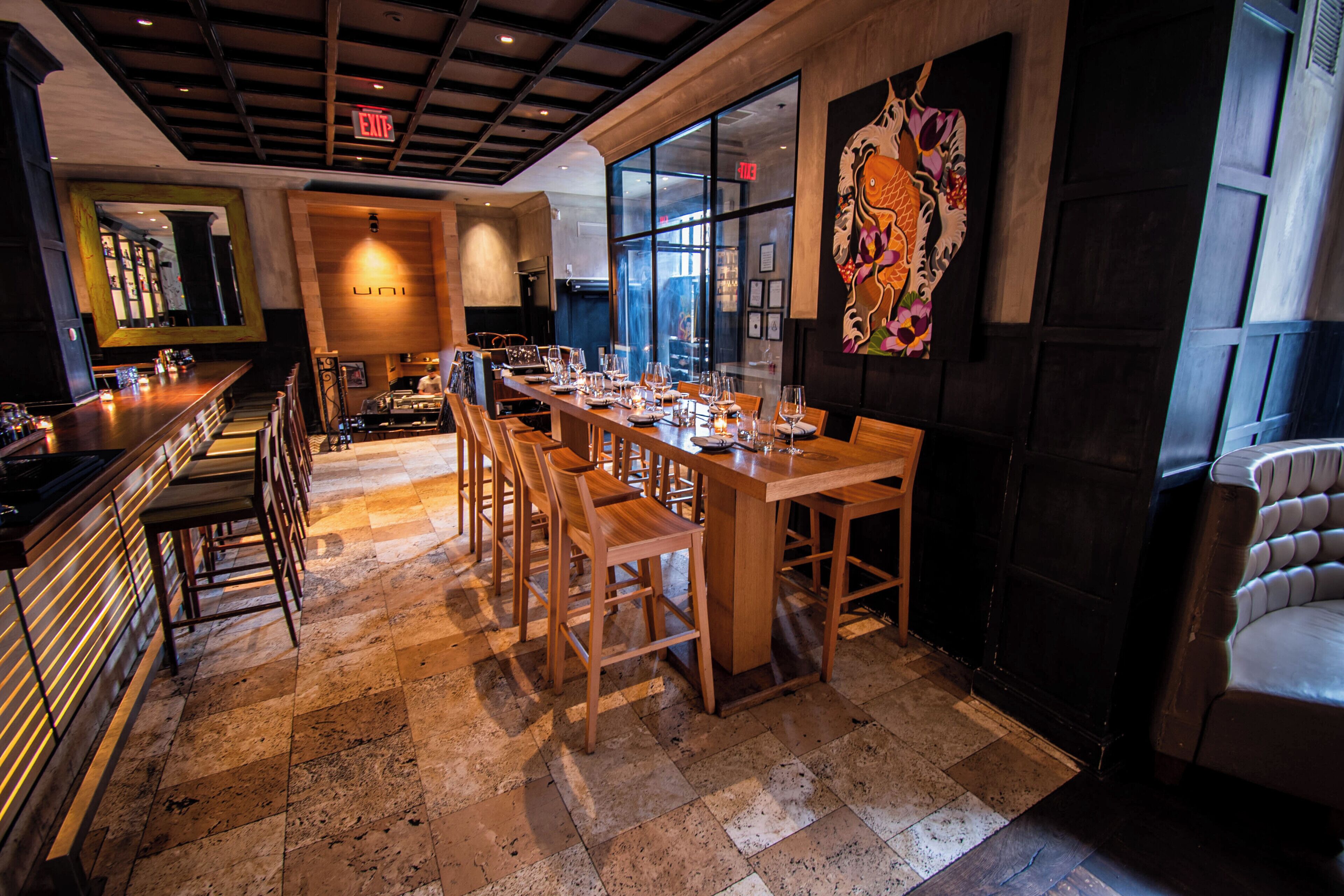 A restaurant with a long wooden table, bar area, and chairs.
