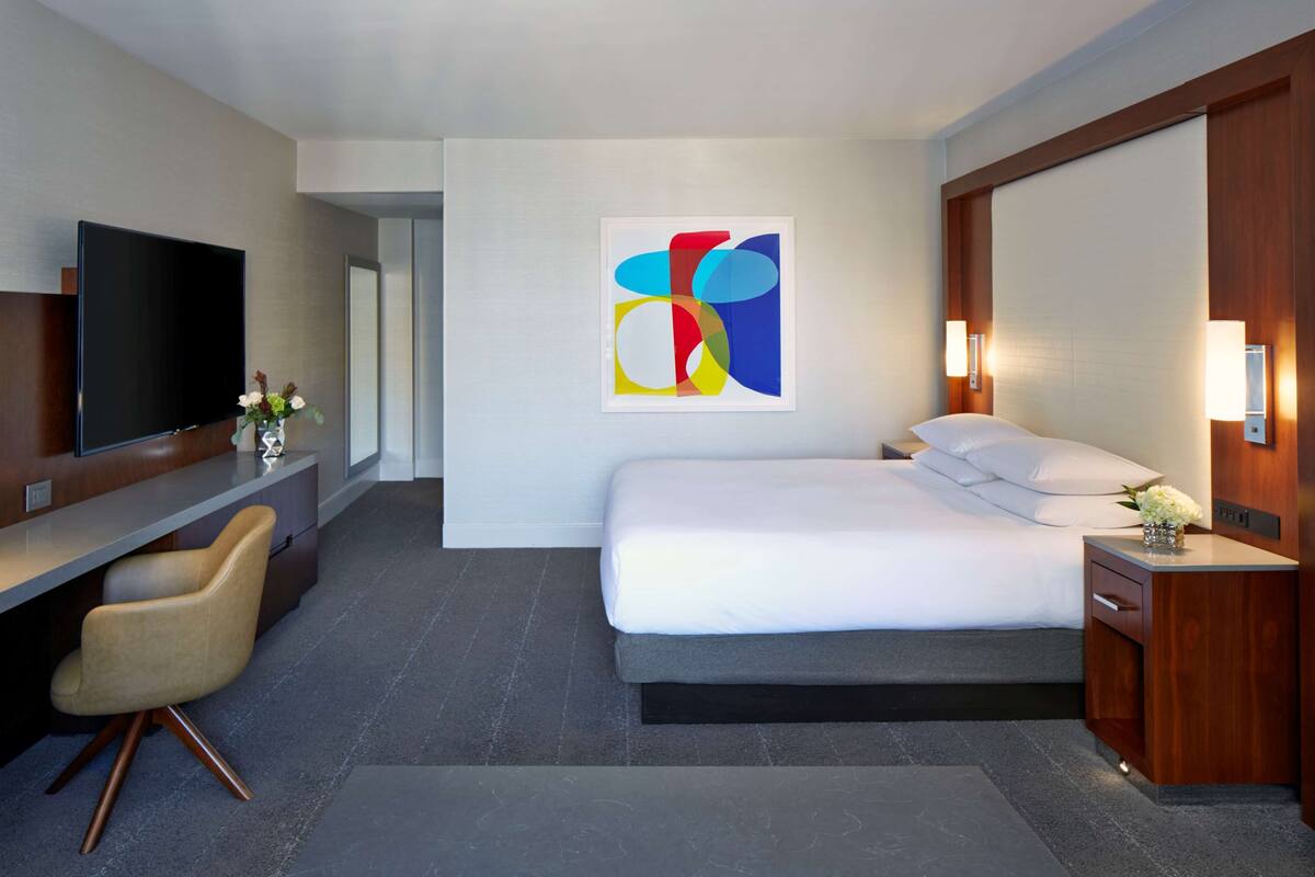 A modern hotel room with a bed, bedside table, TV, and a desk with a chair.