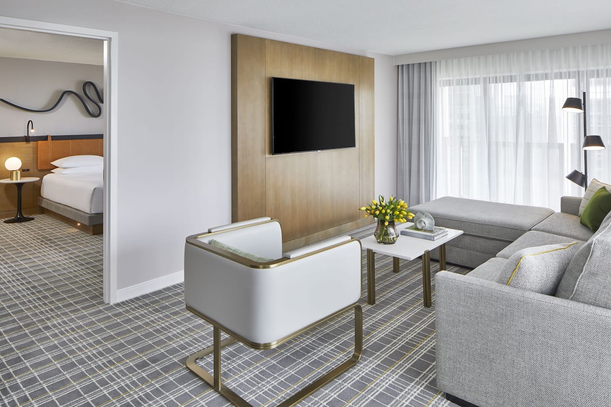 A modern hotel room with a flat-screen TV mounted on a wooden panel, a grey sofa, a white armchair, and a small table with a vase of flowers.