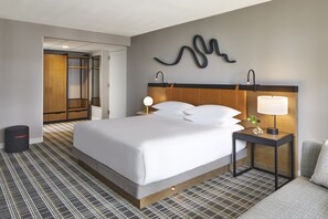 A modern hotel room with a large bed, a bedside table with a lamp, a wall-mounted artwork, and a wardrobe.
