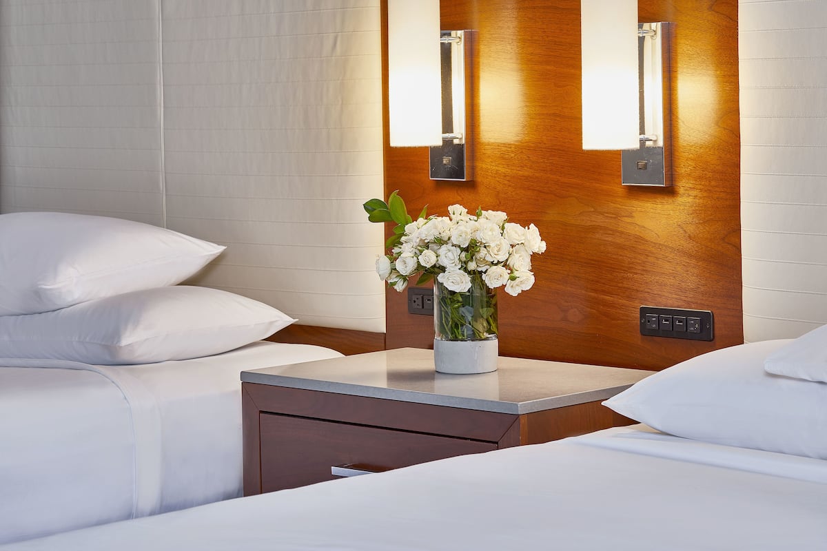 A hotel room with a bed, a bedside table with a vase of white flowers, and wall-mounted light fixtures.