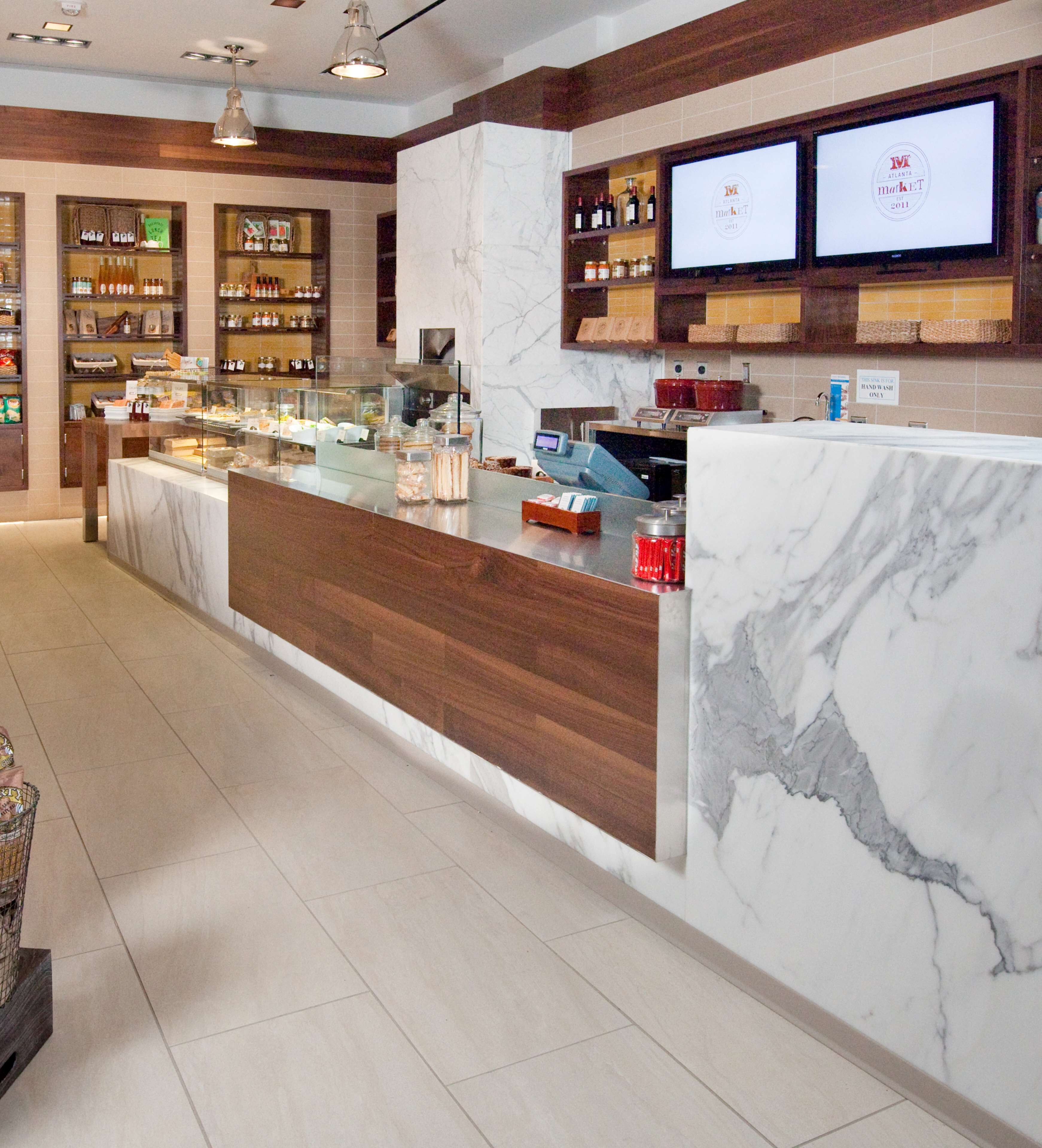 A bakery counter with a marble countertop, wooden shelves with baked goods, and two digital screens displaying the market logo.