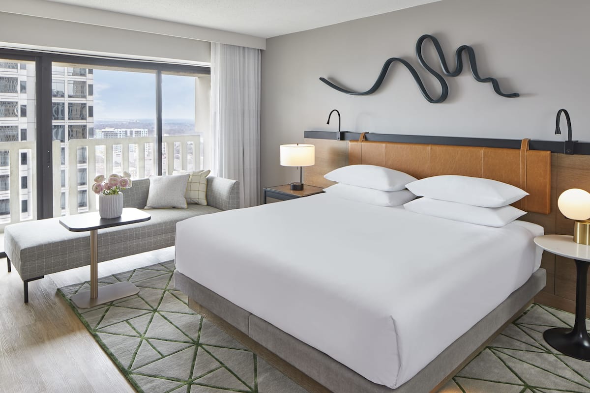 A hotel room with a large bed, a bedside table with a lamp, a small table with a vase of flowers, a sofa, and a view of the city through large windows.