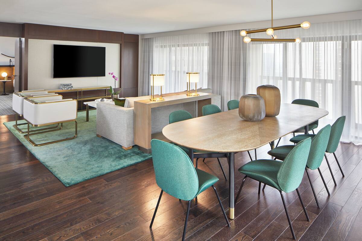 A modern hotel room with a dining area, a flat-screen TV, and a large window with sheer curtains.