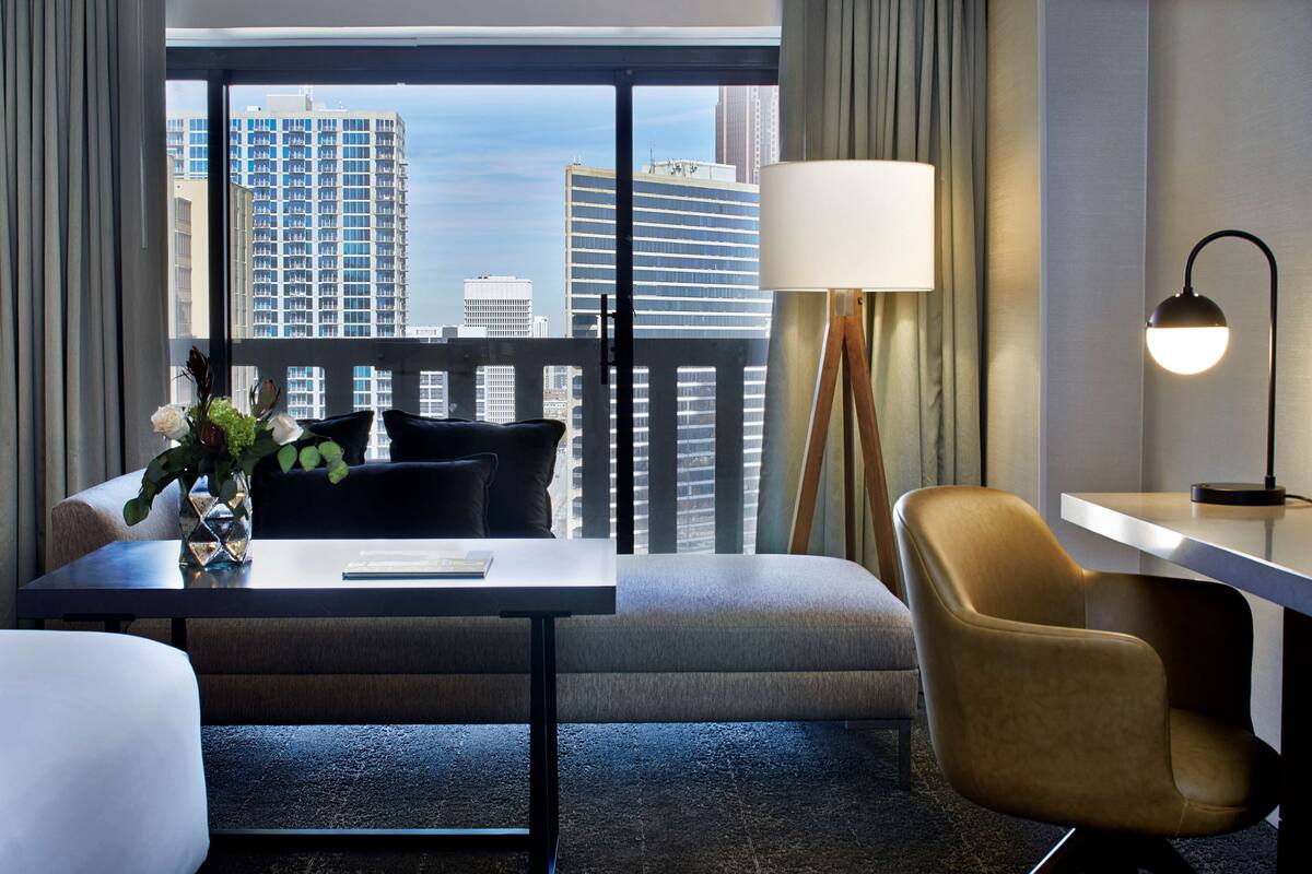 A hotel room with a large window, a desk, a chair, a sofa, and a lamp.
