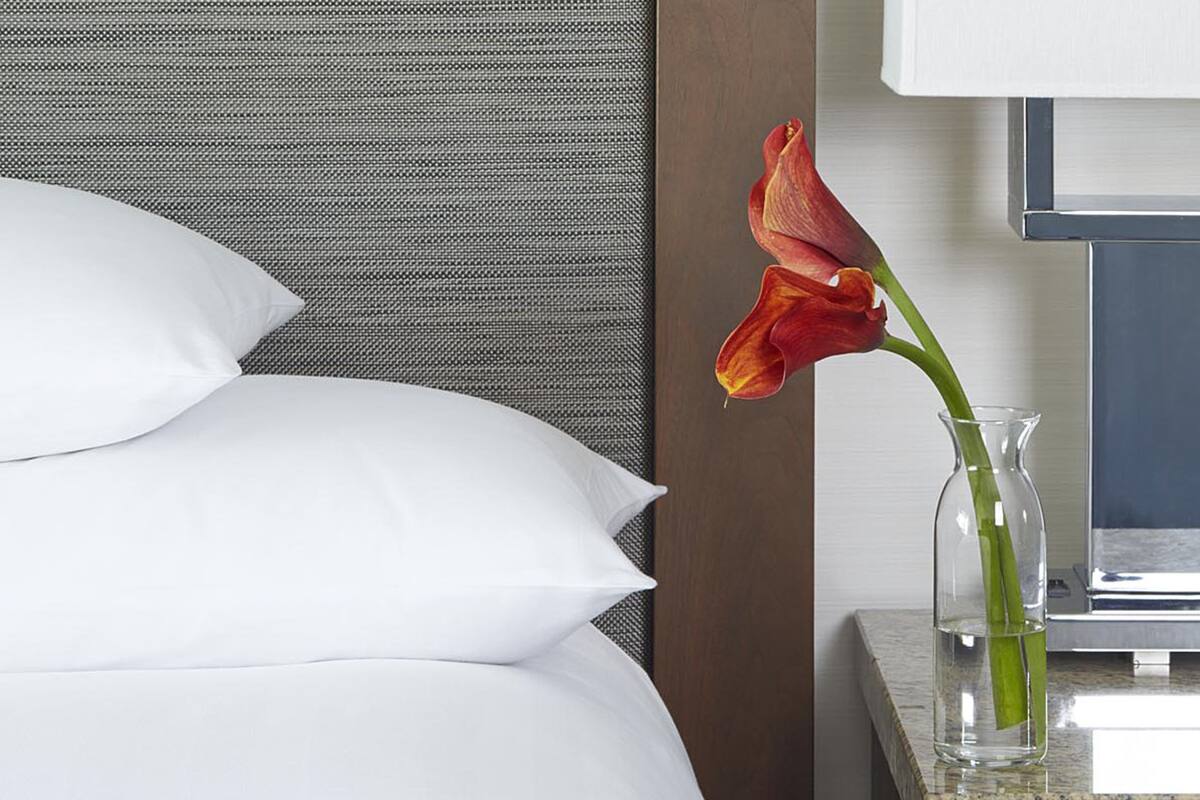 A neatly made bed with white pillows and a vase with a red flower on a bedside table.