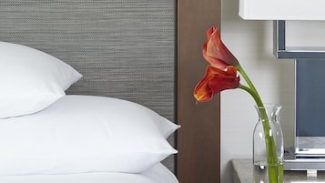 Una cama bien hecha con almohadas blancas y un florero con una flor roja sobre una mesita de noche.