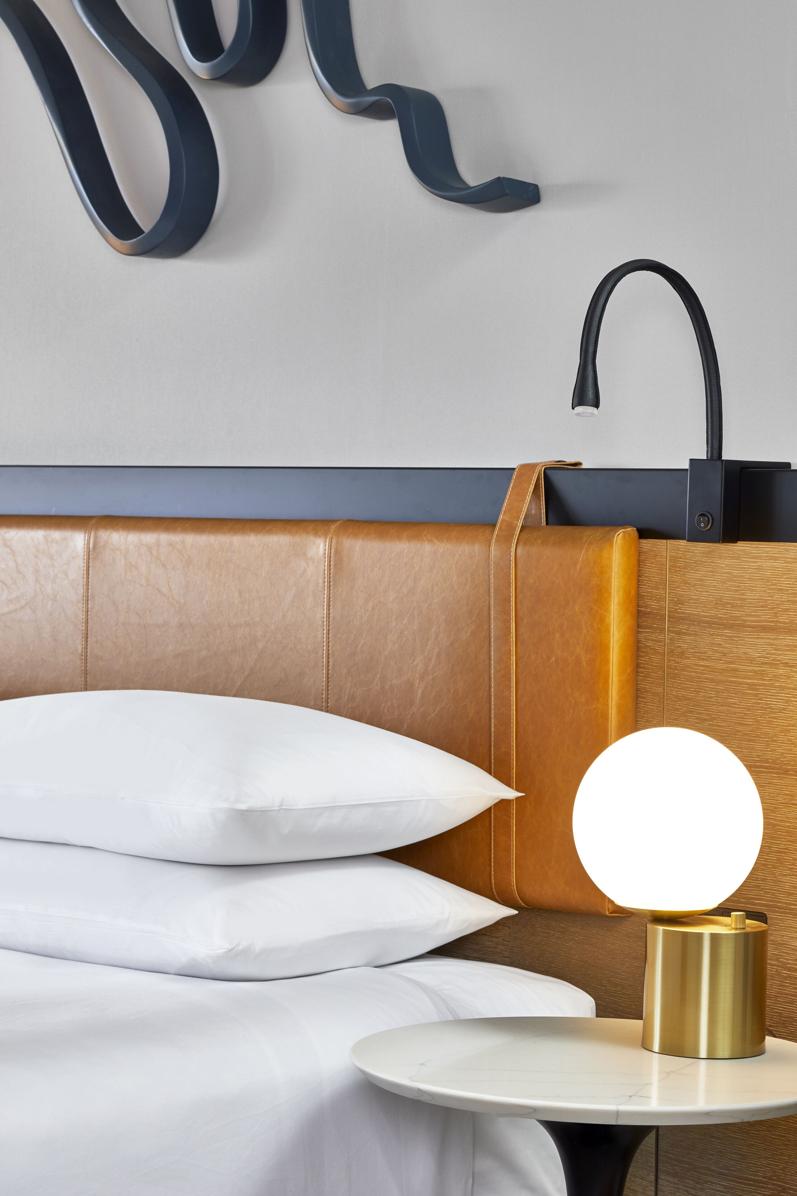 A neatly made bed with white linens, a round gold table lamp, and a wall-mounted black faucet.