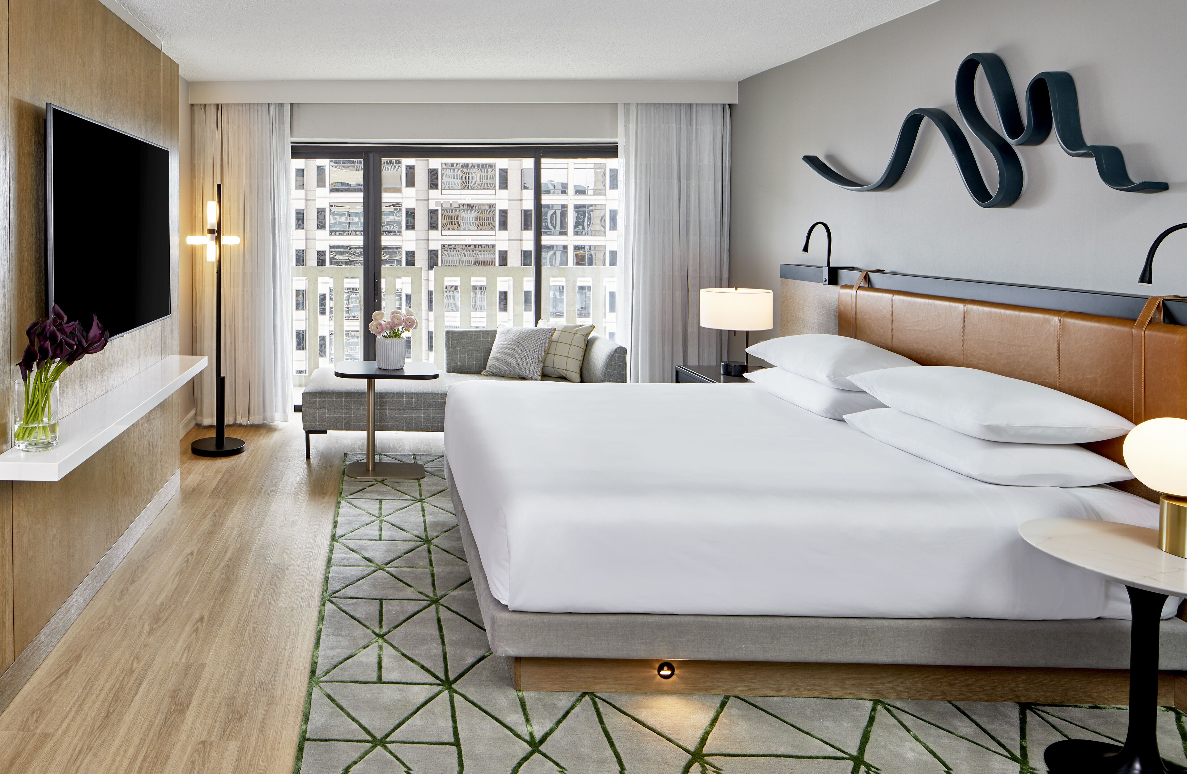 A modern hotel room with a large bed, a TV, a sofa, and a view of the city through large windows.