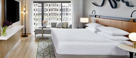A modern hotel room with a large bed, a TV, a sofa, and a view of the city through large windows.