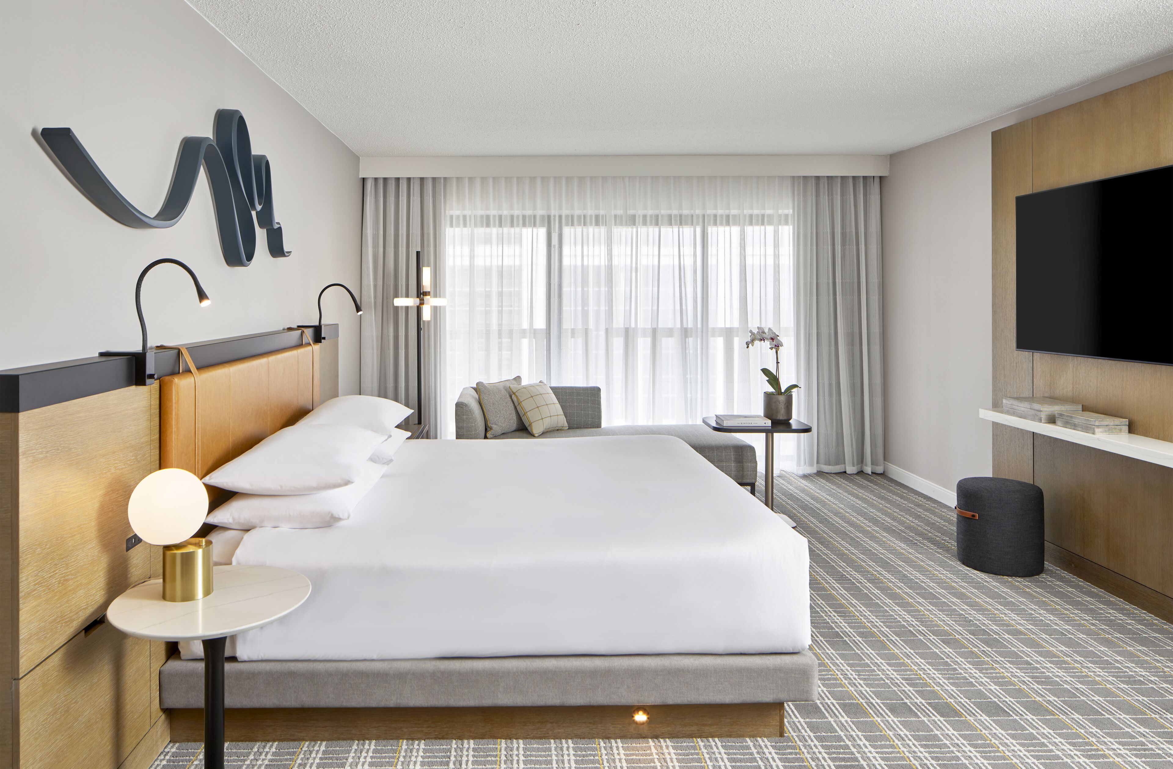 Et moderne hotelværelse med en stor seng, sengeborde med lamper, et fjernsyn, et lille bord med en plante og et vindue med gardiner.