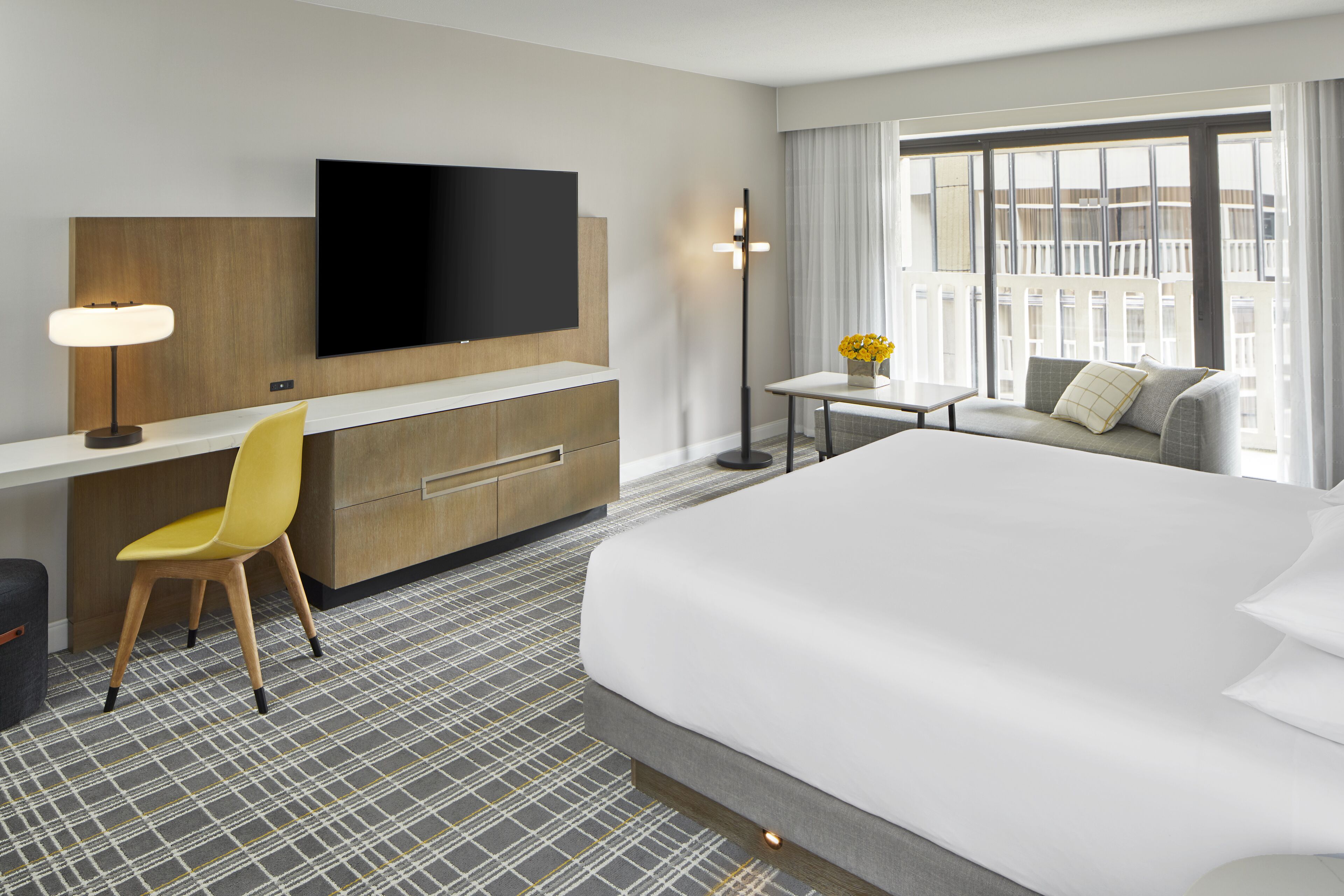A hotel room with a bed, a desk with a chair, a TV mounted on the wall, a small table with a vase of flowers, and a large window with curtains.