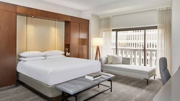 A hotel room with a large bed, a bench, a desk, and a window with a view of buildings.