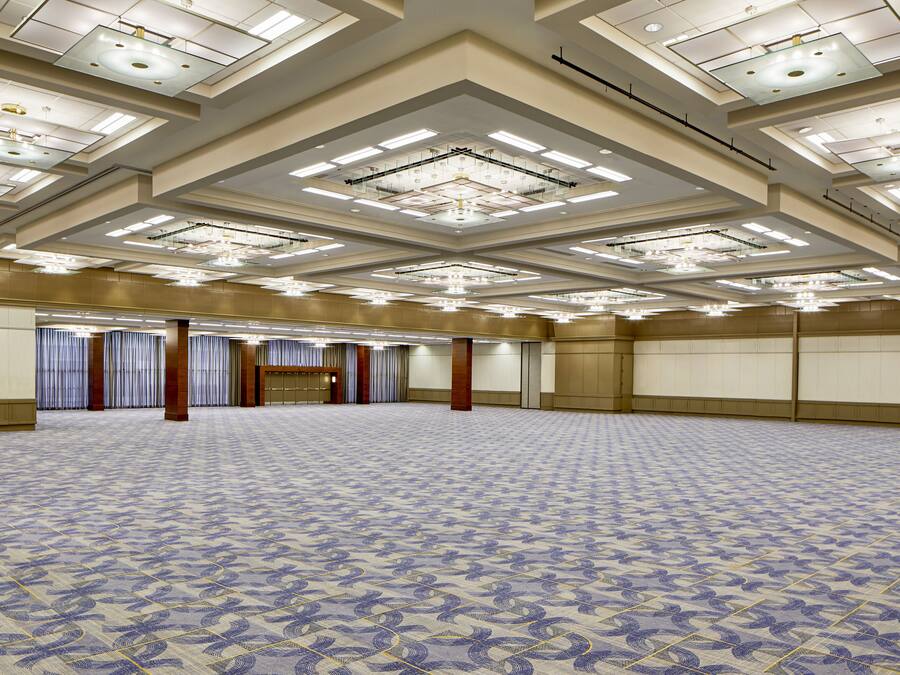 Un salón de conferencias amplio y vacío, con alfombra estampada, techos altos con iluminación empotrada y una paleta de colores neutros.