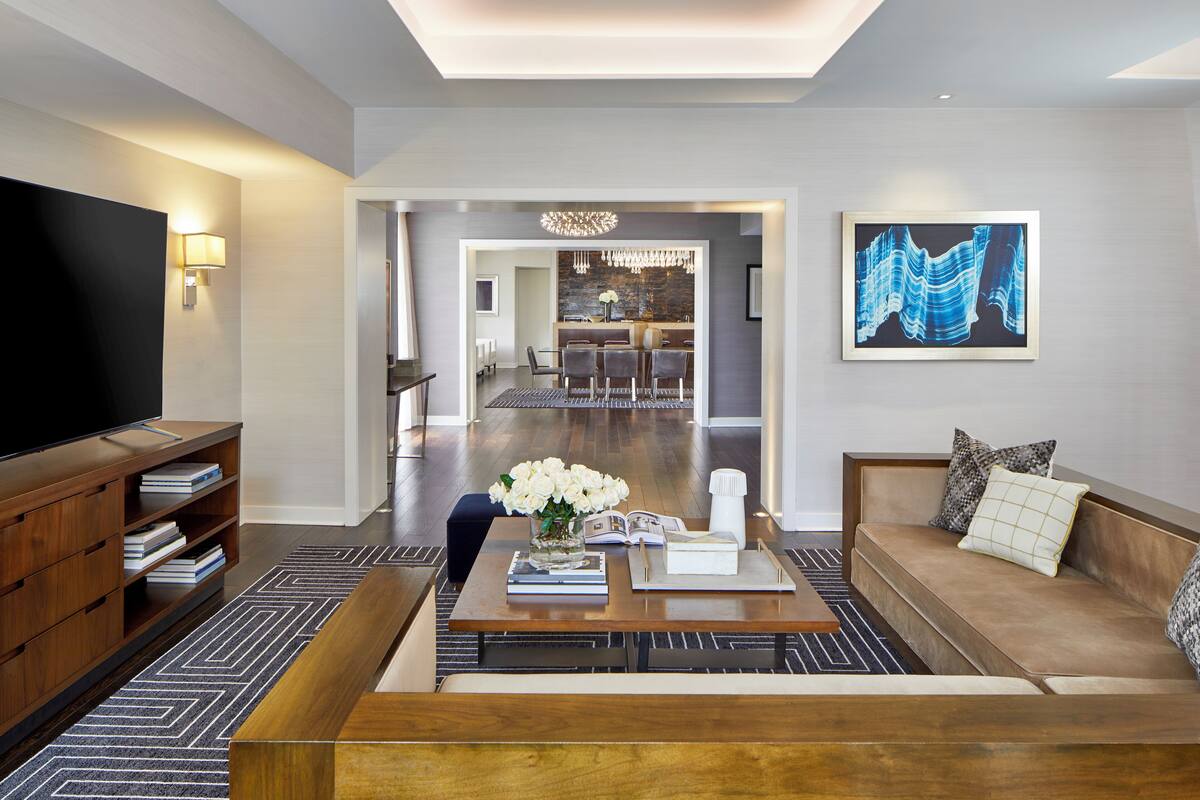 A modern living room with a sofa, coffee table, and TV. There is a dining area in the background and a kitchen visible through an open doorway.