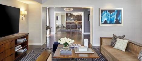 A modern living room with a sofa, coffee table, and TV. There is a dining area in the background and a kitchen visible through an open doorway.