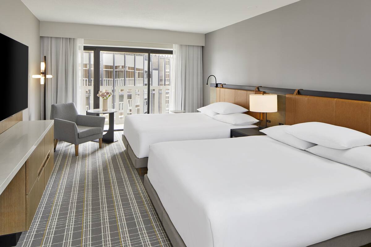 A hotel room with two beds, a desk, a chair, a TV, and a balcony with a view of the city.