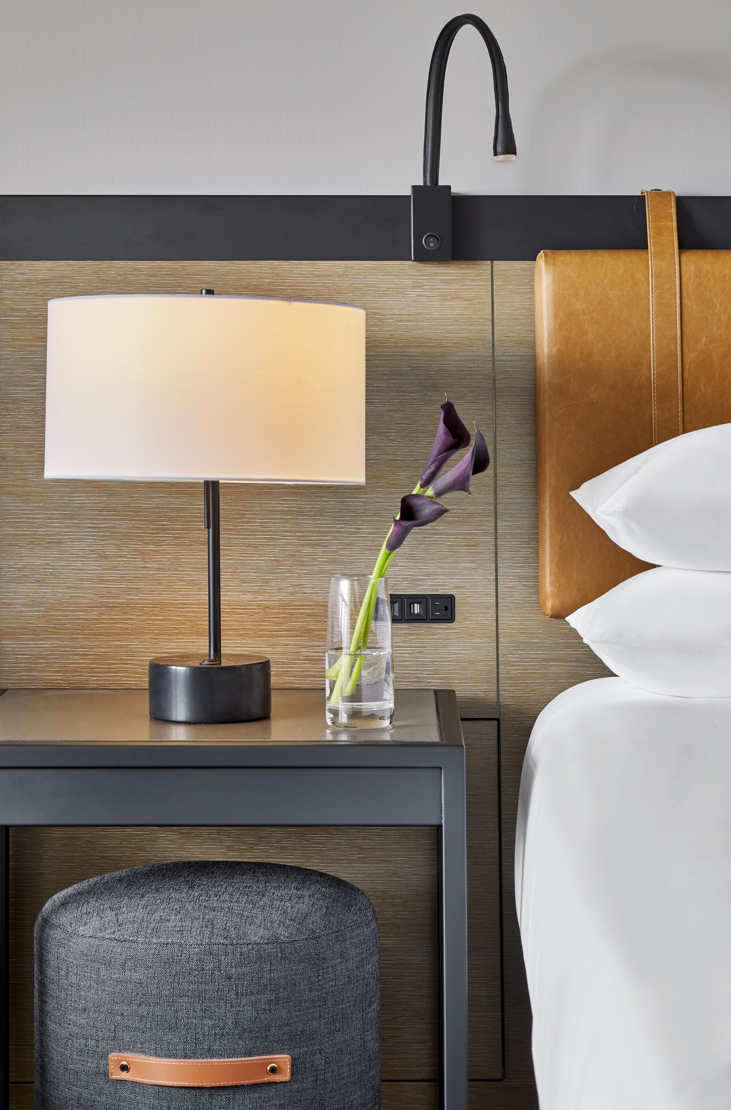 A hotel room with a bed, a desk with a lamp, a vase with flowers, and a modern bathroom design.