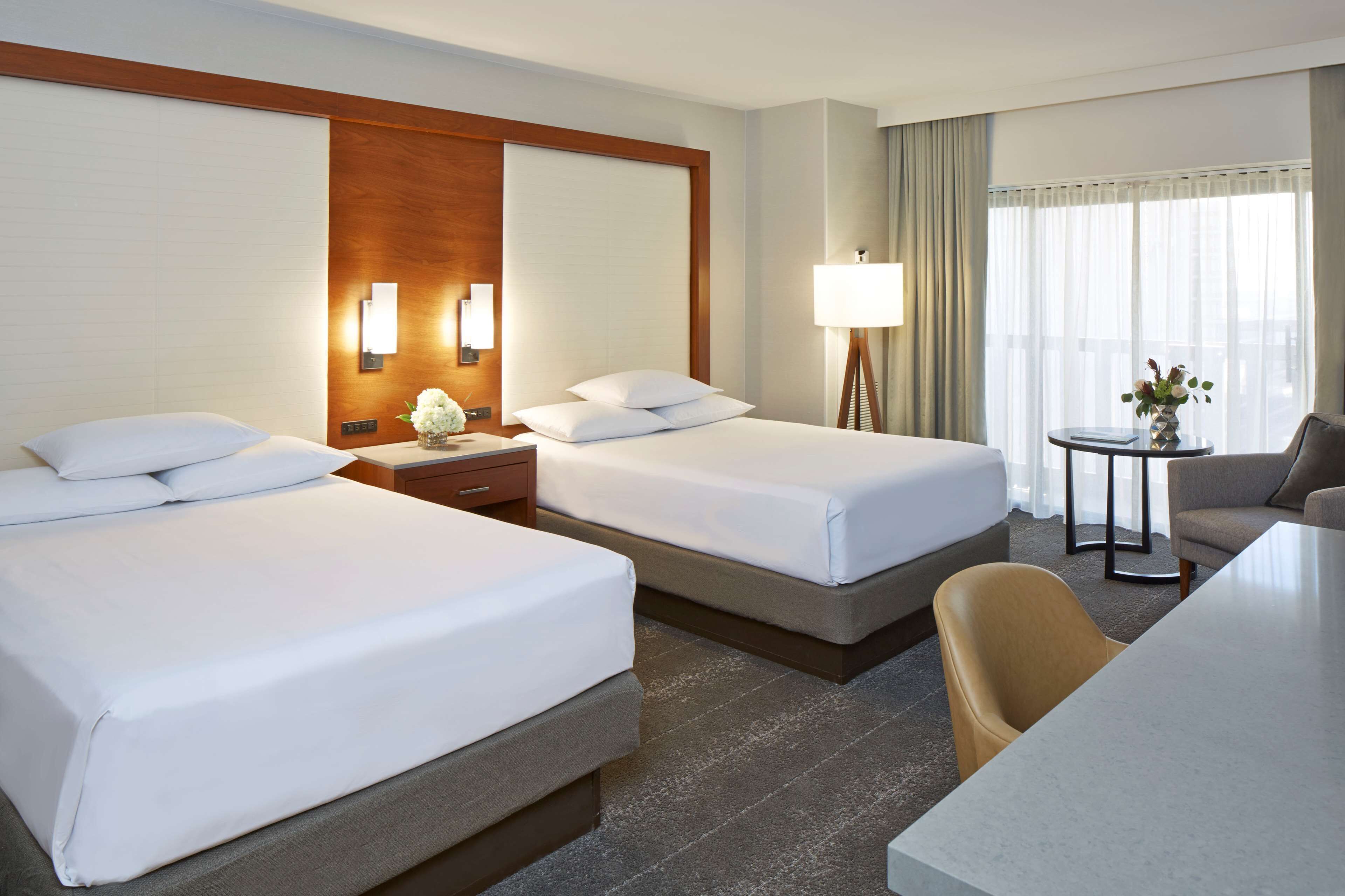 A hotel room with two beds, a desk, a chair, a lamp, and a view of the outdoors.