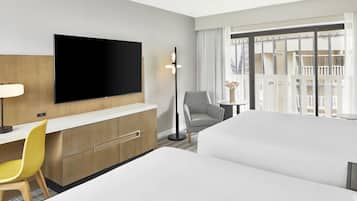 A hotel room with a flat-screen TV, a desk with a chair, a sofa, a small table with a vase, and a large window with curtains.