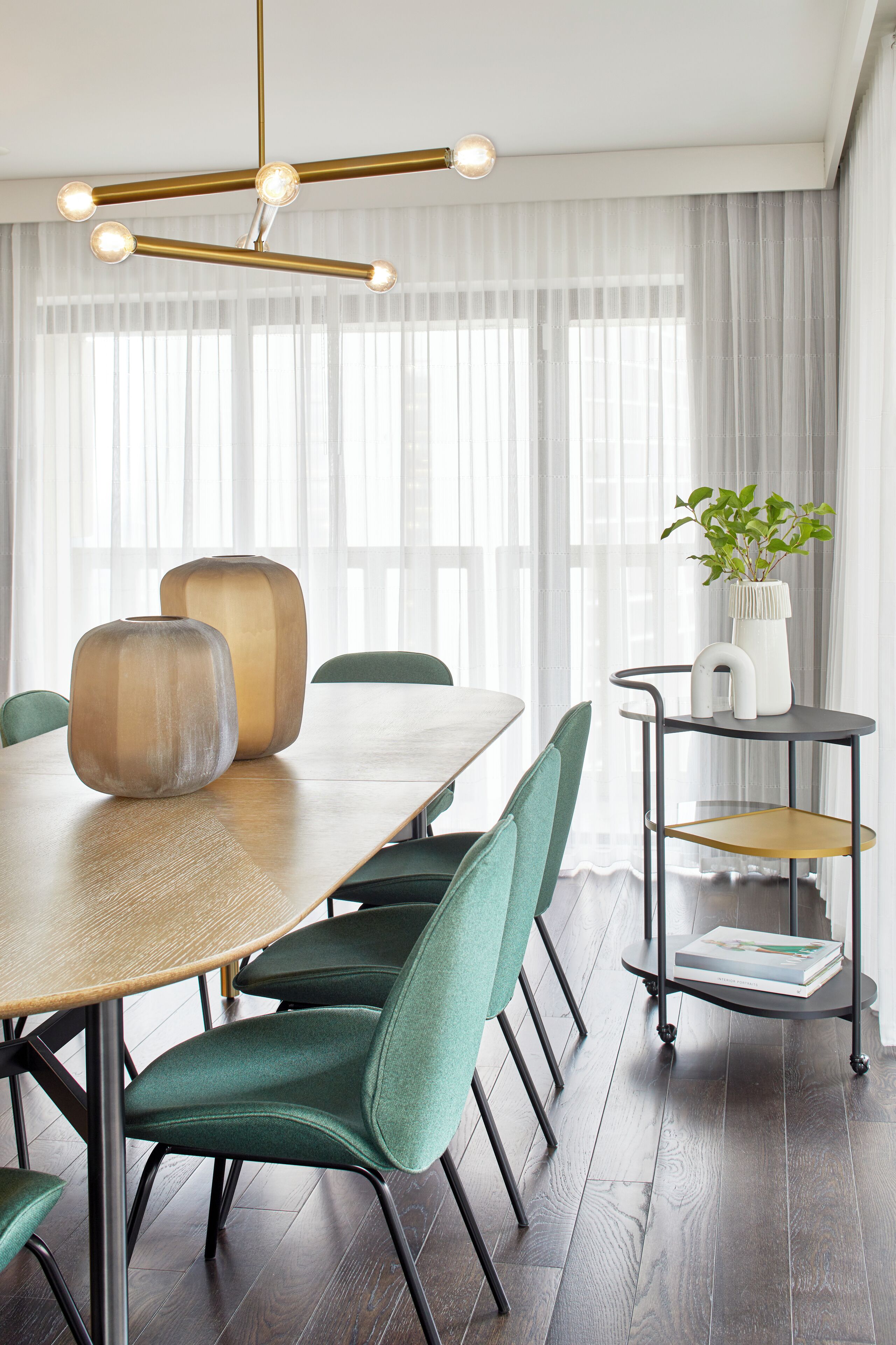 Uma sala de jantar com uma mesa de madeira, cadeiras estofadas em verde, um lustre moderno e uma planta sobre uma mesinha.
