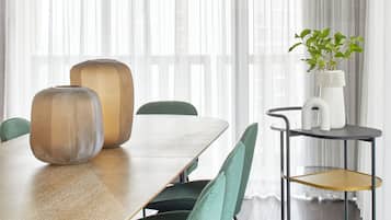 Uma sala de jantar com uma mesa de madeira, cadeiras estofadas em verde, um lustre moderno e uma planta sobre uma mesinha.