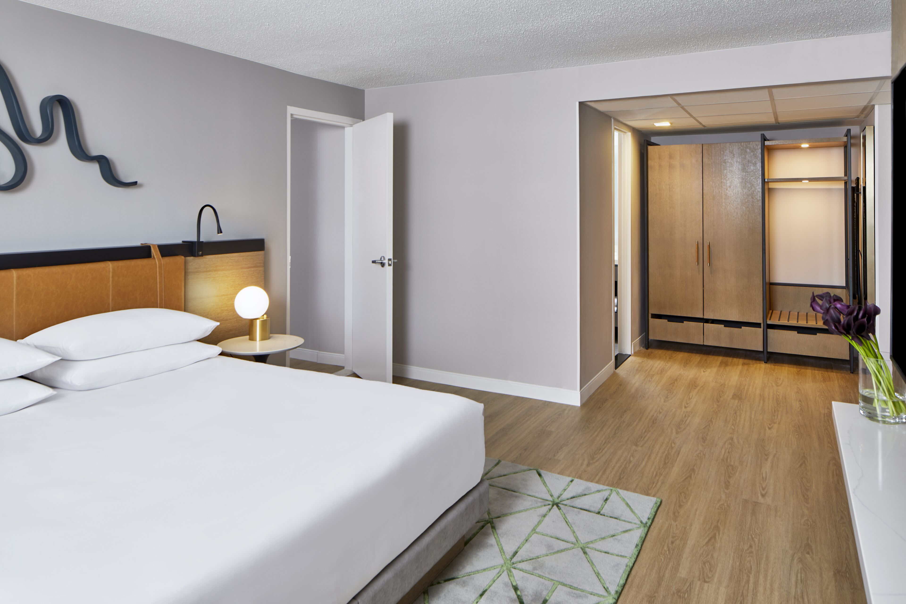 A modern hotel room with a neatly made bed, a bedside lamp, and a view of the hallway leading to a wardrobe.