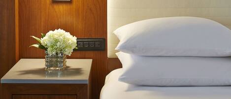 Quarto de hotel com mesa de cabeceira de madeira, mesinha de apoio com um vaso de flores, cama com roupa de cama branca e luminária fixa na parede.