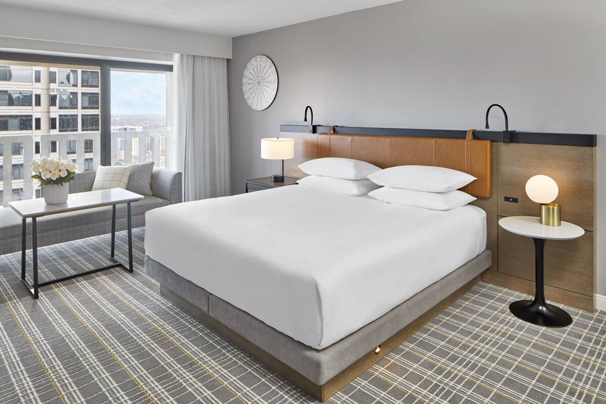 A hotel room with a large bed, a bedside table, a lamp, a clock on the wall, and a view of the city through the window.