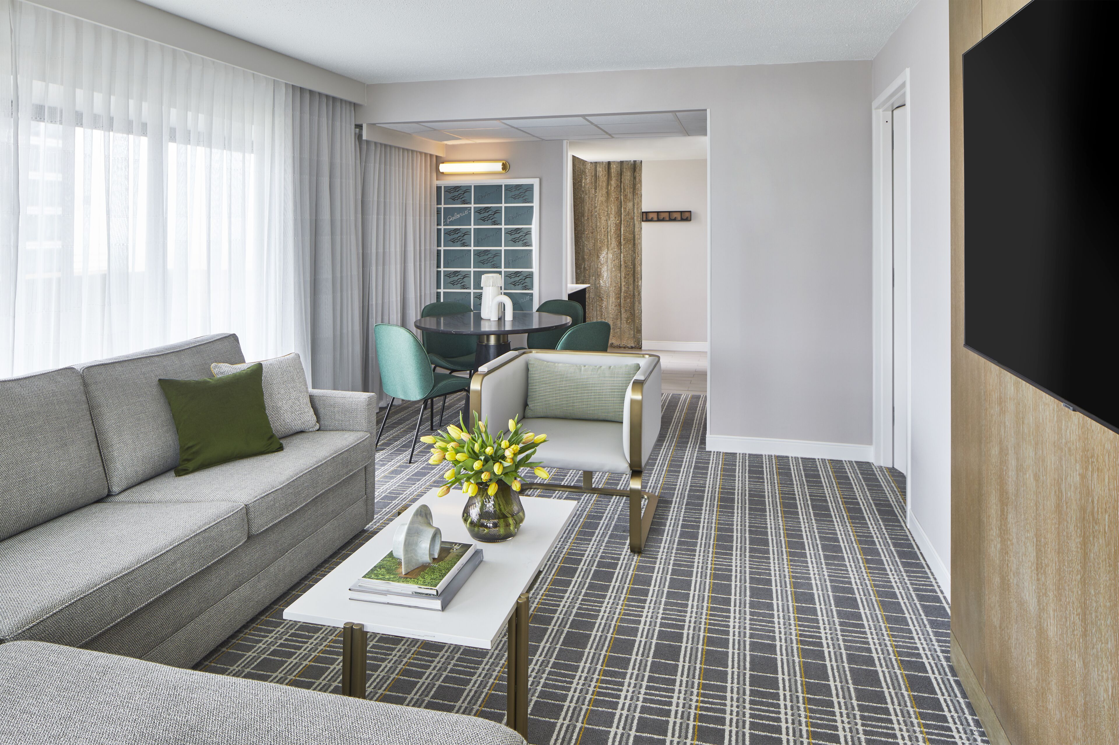 A modern hotel room with a sofa, a dining table with chairs, a coffee table with a vase of flowers, and a large flat-screen TV.