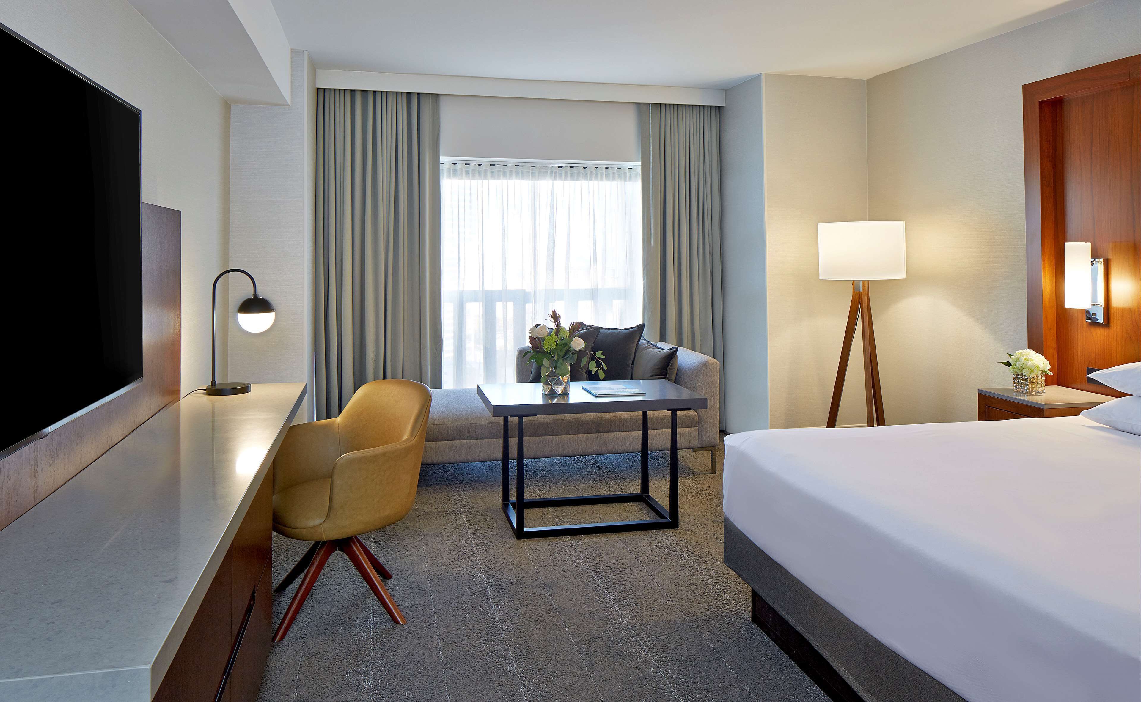 A hotel room with a bed, a desk with a chair, a sofa, a small table with a vase of flowers, a lamp, and a TV.